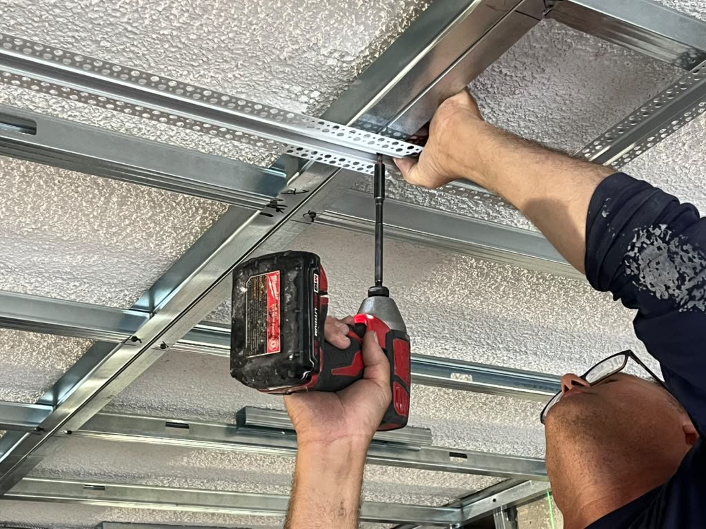 A worker's hands and a drill install a metal ceiling track for a suspended grid system.