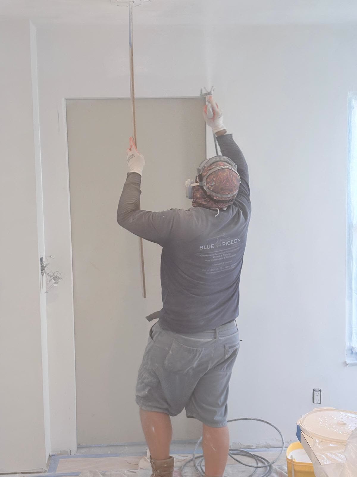 A worker reaches overhead with a pole tool to finish a ceiling corner, wearing a respirator and Blue Pigeon shirt.