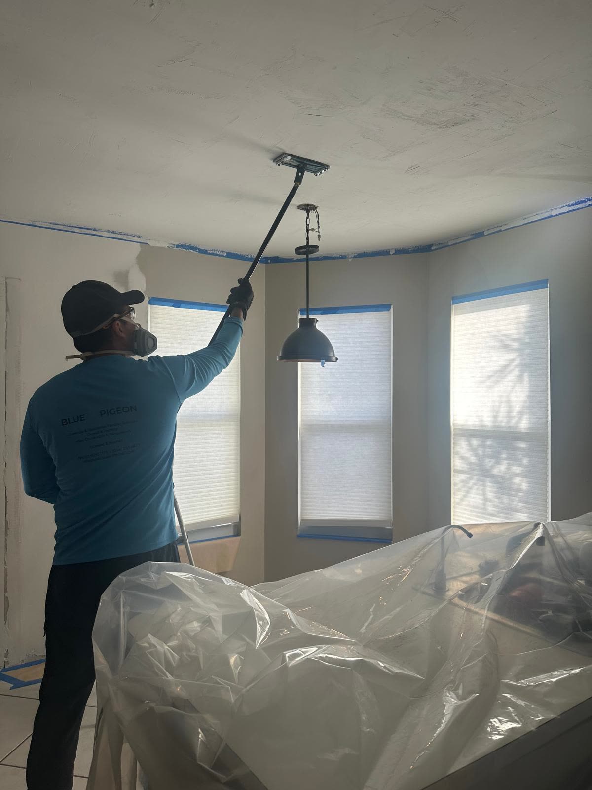 A painter reaches a vaulted ceiling with an extension pole, windows masked and furniture wrapped in plastic.