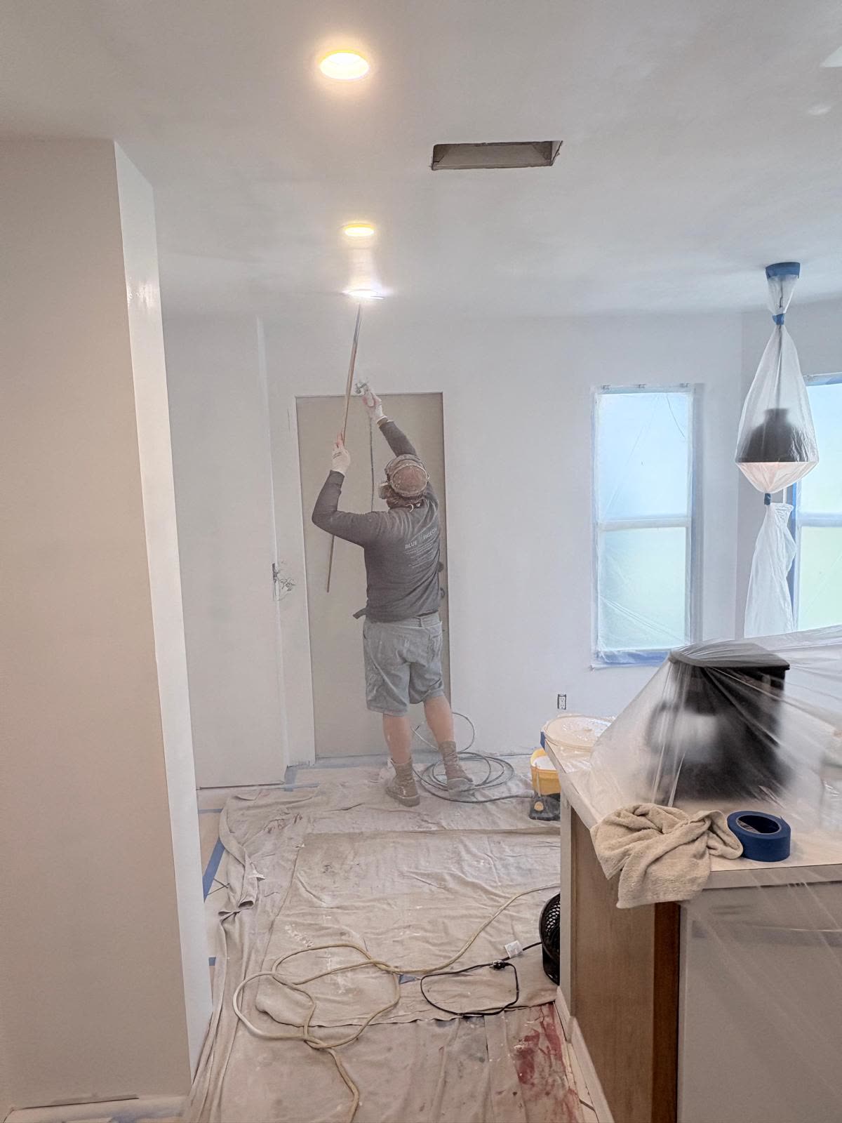 A painter works overhead near recessed lighting in a kitchen protected with plastic sheeting and blue tape.
