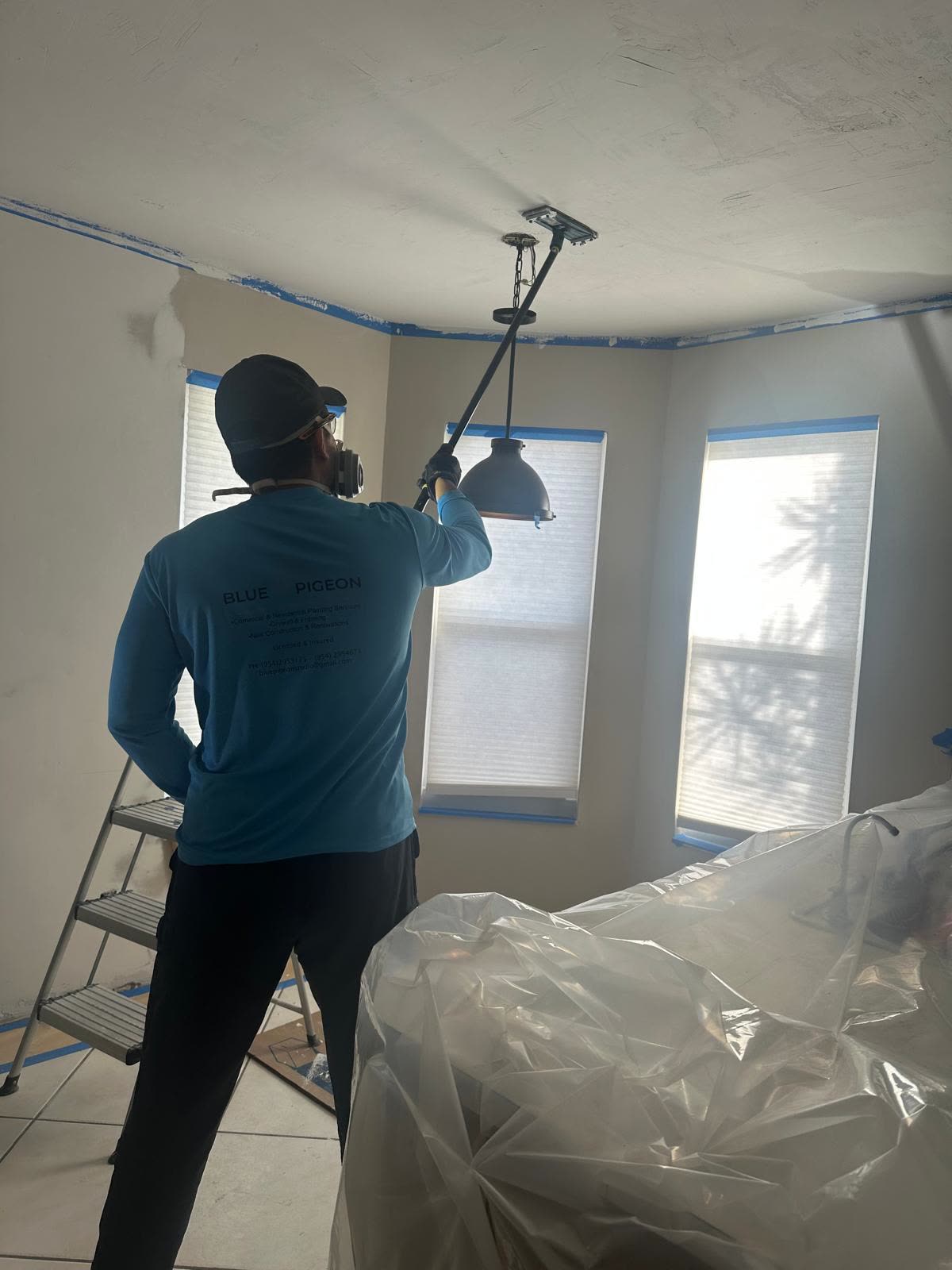 A painter in a respirator reaches up to paint a corner ceiling near a pendant light, protective tape along the trim.